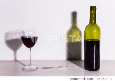 Bottle with wine and glass, red puddle of wine on the table. Cleaning after party concept. Bottle with wine and glass, red puddle of wine on the table. Cleaning after party concept. 65034829
