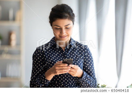 Pleasant millennial indian girl holding smartphone, reading good news sms. Pleasant millennial indian girl holding smartphone, reading good news sms. 65040418