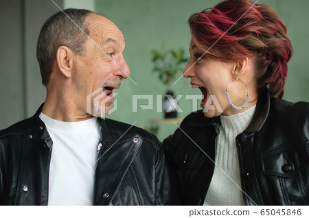 Emotional family portrait of adult daughter and senior father in loft room with houseplants. Screaming man and girl in black leather jackets in punk style and looking to each other face 65045846