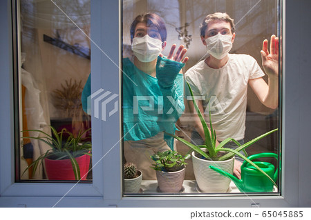 Man and woman with medical mask on their faces looking through window, selfisolation to prevent spread infection. 65045885