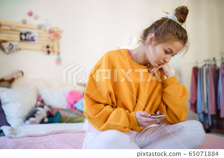 Sad young female student sitting on bed, using smartphone. 65071884