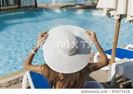 Beautiful woman sunbathing on a beach at tropical travel resort, enjoying summer holidays. Girl holding a hat with her hands on sun lounger near the pool 65073428