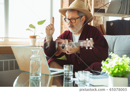 Mature senior older man during quarantine, realizing how important stay at home during virus outbreak, giving concert of taking online lessons of guitar playing 65078003