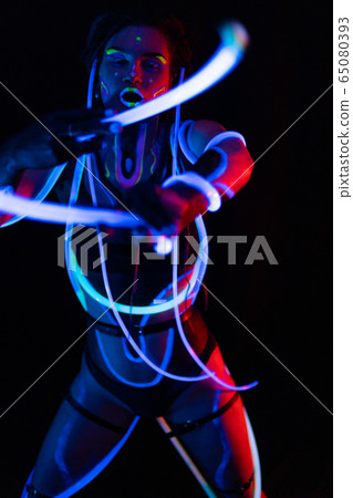 Portrait of a Girl with Glowing Tubes in Neon UF Light. Model Girl with Dreadlocks and Fluorescent Creative Psychedelic MakeUp, Art Design of Female Disco Dancer Model in UV, Colorful Abstract Make-Up 65080393