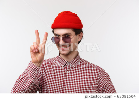 Close-up portrait funny and outgoing hipster man in red beanie, sunglasses and check shirt, getting ready summer vacation, showing peace gesture and smiling, stand white background 65081504