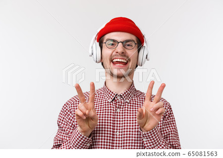 Music, technology and people concept. Positive and happy smiling young man with beard and glasses, wear beanie listen music in headphones, showing peace gesture and laughing, white background 65081563
