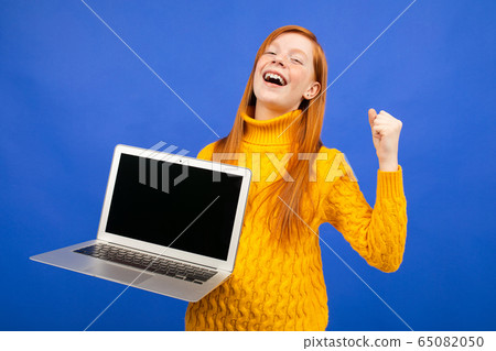 happy red-haired teenager shows a laptop display with a layout for advertising on a blue studio happy red-haired teenager shows a laptop display with a layout for advertising on a blue studio 65082050
