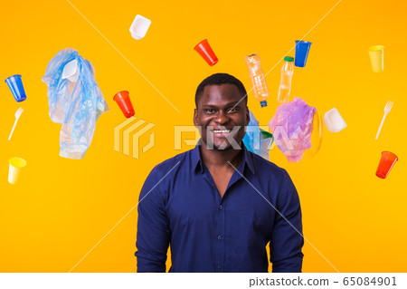 Problem of trash, plastic recycling, pollution and environmental concept - african american man is standing under trash on yellow background Problem of trash, plastic recycling, pollution and environmental concept - african american man is standing under trash on yellow background 65084901