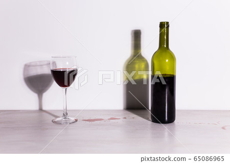 Bottle with wine and glass, red puddle of wine on the table. Cleaning after party concept. Bottle with wine and glass, red puddle of wine on the table. Cleaning after party concept. 65086965