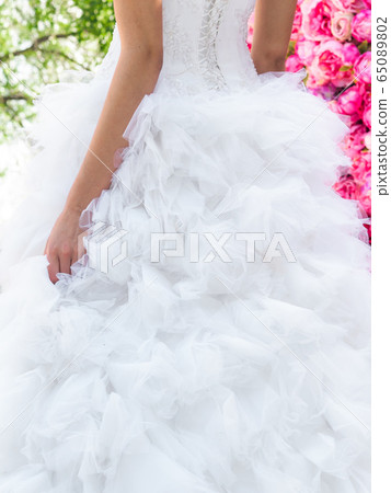 Beautiful fluffy skirt of wedding dress on bride which is posing on a decorative background of pink flowers Beautiful fluffy skirt of wedding dress on bride which is posing on a decorative background of pink flowers 65089802