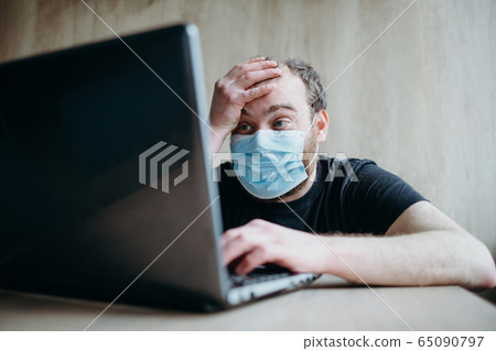 Coronavirus, an adult male with a beard works at a laptop. Protective respiratory mask. Coronavirus, an adult male with a beard works at a laptop. Protective respiratory mask. 65090797