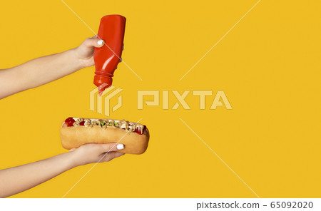 Close up of young woman squeezing ketchup onto hot dog against orange background, copy space. Panorama Close up of young woman squeezing ketchup onto hot dog against orange background, copy space. Panorama 65092020