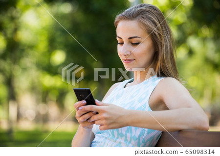 girl sitting on a bench and using phone in park 65098314