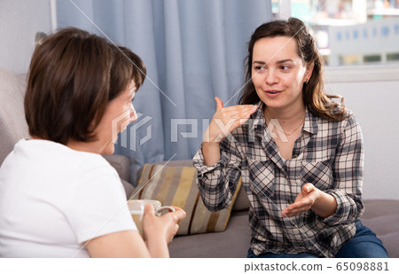 Smiling young female friends gossiping on sofa in home interior Smiling young female friends gossiping on sofa in home interior 65098881
