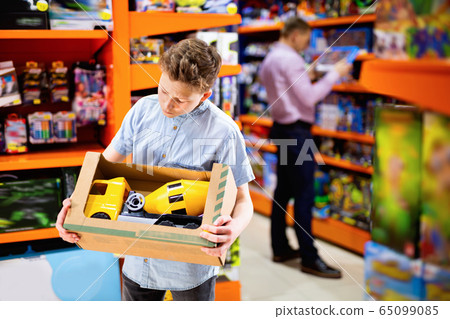 Serious teen boy looking narrowly at yellow plastic car in children toy store 65099085