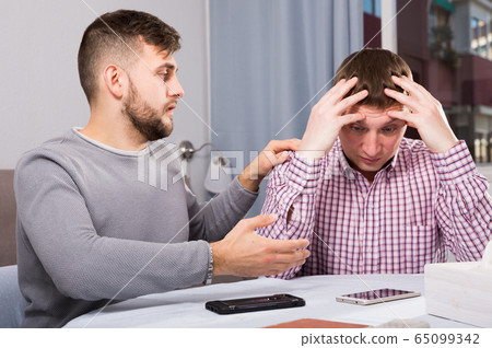 Friend soothing upset man at home table 65099342
