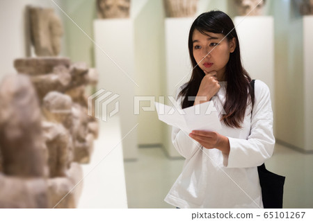 Chinese woman visitor with guide book looking at exhibition in museum 65101267