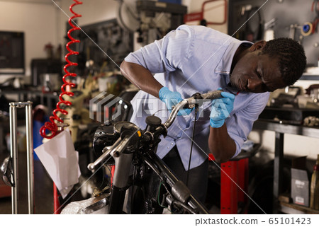 Confident afro american worker using his tools for repairing in motorcycle workshop Confident afro american worker using his tools for repairing in motorcycle workshop 65101423