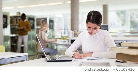 Portrait of smiling woman with laptop and book in public library Portrait of smiling woman with laptop and book in public library 65102436