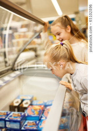 Two small sisters standing near fridge choosing frozen food Two small sisters standing near fridge choosing frozen food 65102896