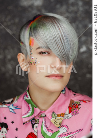 Portrait of a young beautiful girl in a pink shirt on a gray background with dyed hair. Short haircut pixie or bob. Rainbow coloring and sequins on the face. 65108931