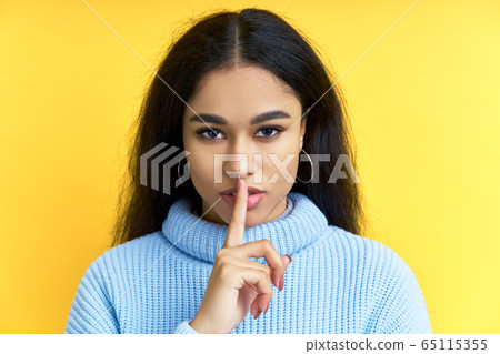Pretty black woman keeps finger on lips, making hush gesture on yellow background 65115355