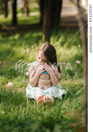 Cute little girl sits on the grass barefoot and hug teddy bear. Kid spend time in the park, outside. Beautiful little girl model 65120597