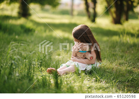 Cute little girl sits on the grass barefoot and hug teddy bear. Kid spend time in the park, outside. Beautiful little girl model Cute little girl sits on the grass barefoot and hug teddy bear. Kid spend time in the park, outside. Beautiful little girl model 65120598