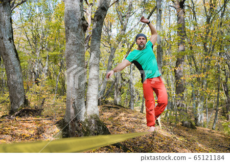 An aged bearded man in orange pants walks on a slack stretched between trees in an autumn forest. The concept of balance and sports lifestyle for people over 40 An aged bearded man in orange pants walks on a slack stretched between trees in an autumn forest. The concept of balance and sports lifestyle for people over 40 65121184