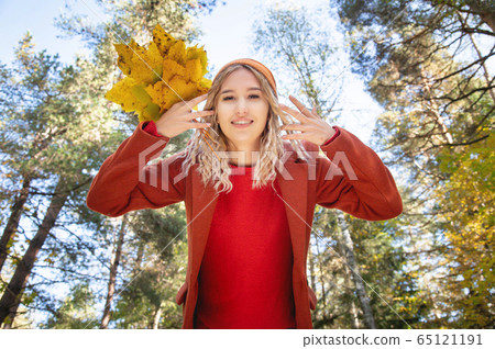female portrait. Beautiful blonde girl in a black felt hat and a brown knitted sweater hides her face behind a yellow maple leaf. horizontal autumn photo 65121191