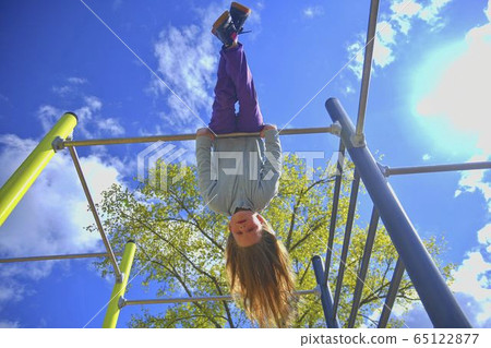 Beautiful blond girl with long hair playing in playground. young girl hanging upside down in a park. 65122877