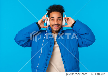 Waist-up portrait happy smiling african american guy listening music in headphones, put earphones as walking outside on winter holidays, wearing padded jacket, standing blue background 65130304