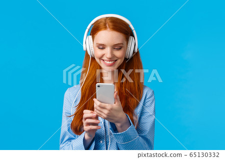 Technology addication, lifestyle and women concept. Cheerful good-looking redhead woman in nightwear, wearing big headphones listen music, smiling pick songs in smartphone app 65130332