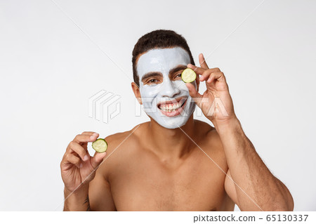 Photo of joyful african american man in facial cosmetic mask making fun with cucumber isolated over white background 65130337
