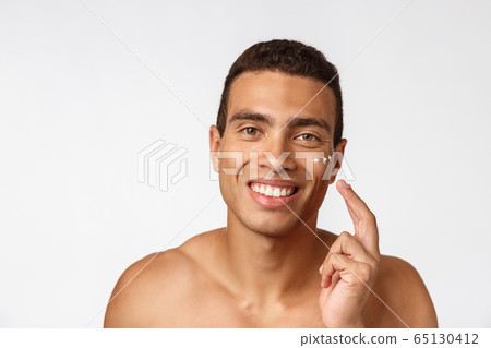 Photo of shirtless african american man smiling and applying face cream isolated over white background 65130412