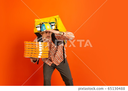 Contacless delivery service during quarantine. Man delivers food and shopping bags during insulation. Emotions of deliveryman isolated on orange background. Contacless delivery service during quarantine. Man delivers food and shopping bags during insulation. Emotions of deliveryman isolated on orange background. 65130488