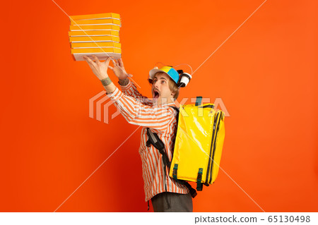 Contacless delivery service during quarantine. Man delivers food and shopping bags during insulation. Emotions of deliveryman isolated on orange background. Contacless delivery service during quarantine. Man delivers food and shopping bags during insulation. Emotions of deliveryman isolated on orange background. 65130498