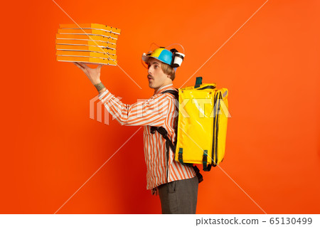 Contacless delivery service during quarantine. Man delivers food and shopping bags during insulation. Emotions of deliveryman isolated on orange background. Contacless delivery service during quarantine. Man delivers food and shopping bags during insulation. Emotions of deliveryman isolated on orange background. 65130499
