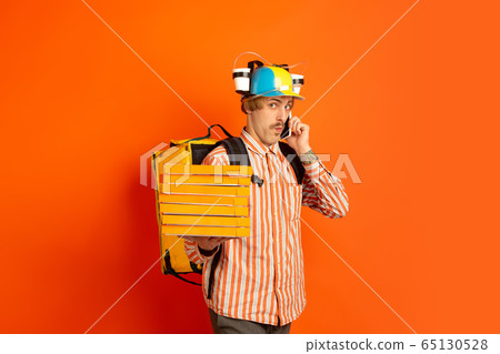 Contacless delivery service during quarantine. Man delivers food and shopping bags during insulation. Emotions of deliveryman isolated on orange background. 65130528