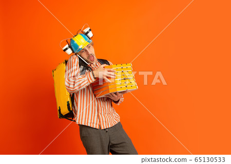 Contacless delivery service during quarantine. Man delivers food and shopping bags during insulation. Emotions of deliveryman isolated on orange background. Contacless delivery service during quarantine. Man delivers food and shopping bags during insulation. Emotions of deliveryman isolated on orange background. 65130533