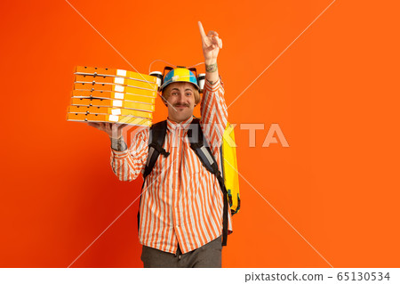 Contacless delivery service during quarantine. Man delivers food and shopping bags during insulation. Emotions of deliveryman isolated on orange background. Contacless delivery service during quarantine. Man delivers food and shopping bags during insulation. Emotions of deliveryman isolated on orange background. 65130534