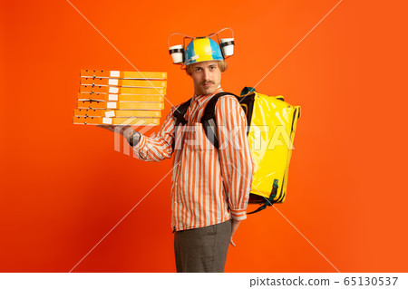 Contacless delivery service during quarantine. Man delivers food and shopping bags during insulation. Emotions of deliveryman isolated on orange background. Contacless delivery service during quarantine. Man delivers food and shopping bags during insulation. Emotions of deliveryman isolated on orange background. 65130537