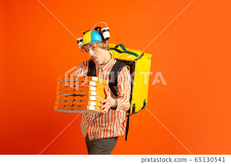 Contacless delivery service during quarantine. Man delivers food and shopping bags during insulation. Emotions of deliveryman isolated on orange background. Contacless delivery service during quarantine. Man delivers food and shopping bags during insulation. Emotions of deliveryman isolated on orange background. 65130541