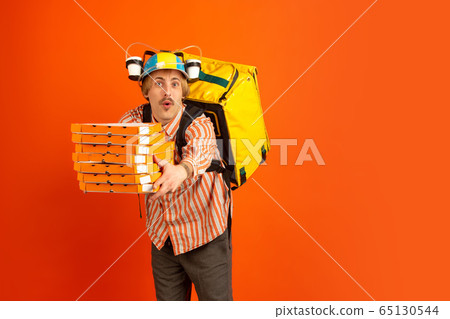 Contacless delivery service during quarantine. Man delivers food and shopping bags during insulation. Emotions of deliveryman isolated on orange background. Contacless delivery service during quarantine. Man delivers food and shopping bags during insulation. Emotions of deliveryman isolated on orange background. 65130544
