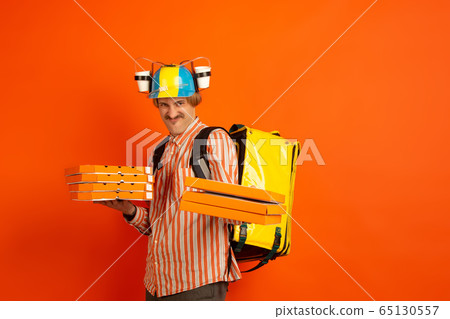 Contacless delivery service during quarantine. Man delivers food and shopping bags during insulation. Emotions of deliveryman isolated on orange background. Contacless delivery service during quarantine. Man delivers food and shopping bags during insulation. Emotions of deliveryman isolated on orange background. 65130557