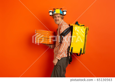 Contacless delivery service during quarantine. Man delivers food and shopping bags during insulation. Emotions of deliveryman isolated on orange background. Contacless delivery service during quarantine. Man delivers food and shopping bags during insulation. Emotions of deliveryman isolated on orange background. 65130563
