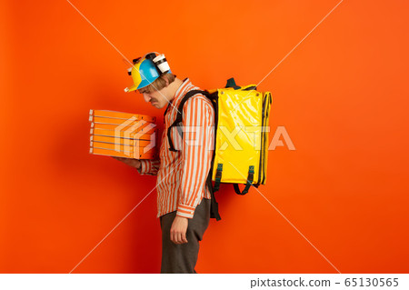 Contacless delivery service during quarantine. Man delivers food and shopping bags during insulation. Emotions of deliveryman isolated on orange background. Contacless delivery service during quarantine. Man delivers food and shopping bags during insulation. Emotions of deliveryman isolated on orange background. 65130565
