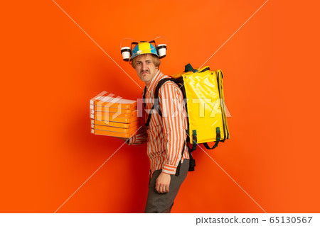 Contacless delivery service during quarantine. Man delivers food and shopping bags during insulation. Emotions of deliveryman isolated on orange background. Contacless delivery service during quarantine. Man delivers food and shopping bags during insulation. Emotions of deliveryman isolated on orange background. 65130567
