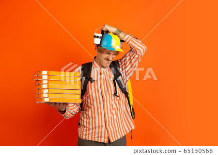 Contacless delivery service during quarantine. Man delivers food and shopping bags during insulation. Emotions of deliveryman isolated on orange background. 65130568