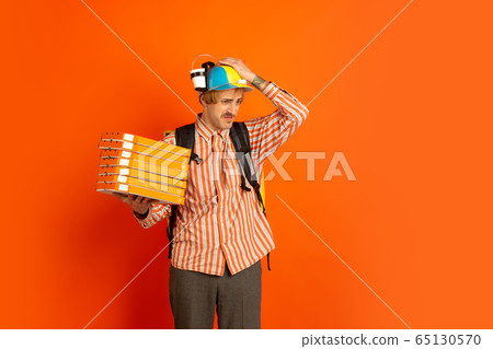 Contacless delivery service during quarantine. Man delivers food and shopping bags during insulation. Emotions of deliveryman isolated on orange background. Contacless delivery service during quarantine. Man delivers food and shopping bags during insulation. Emotions of deliveryman isolated on orange background. 65130570